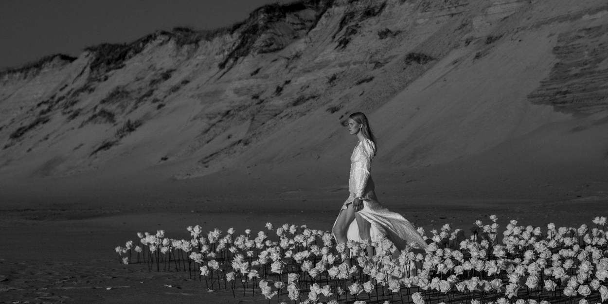 Black-and-white bridal editorial by a Washington DC photographer: a bride in a flowing white gown walks through a field of white roses on a beach beneath a sand dune.