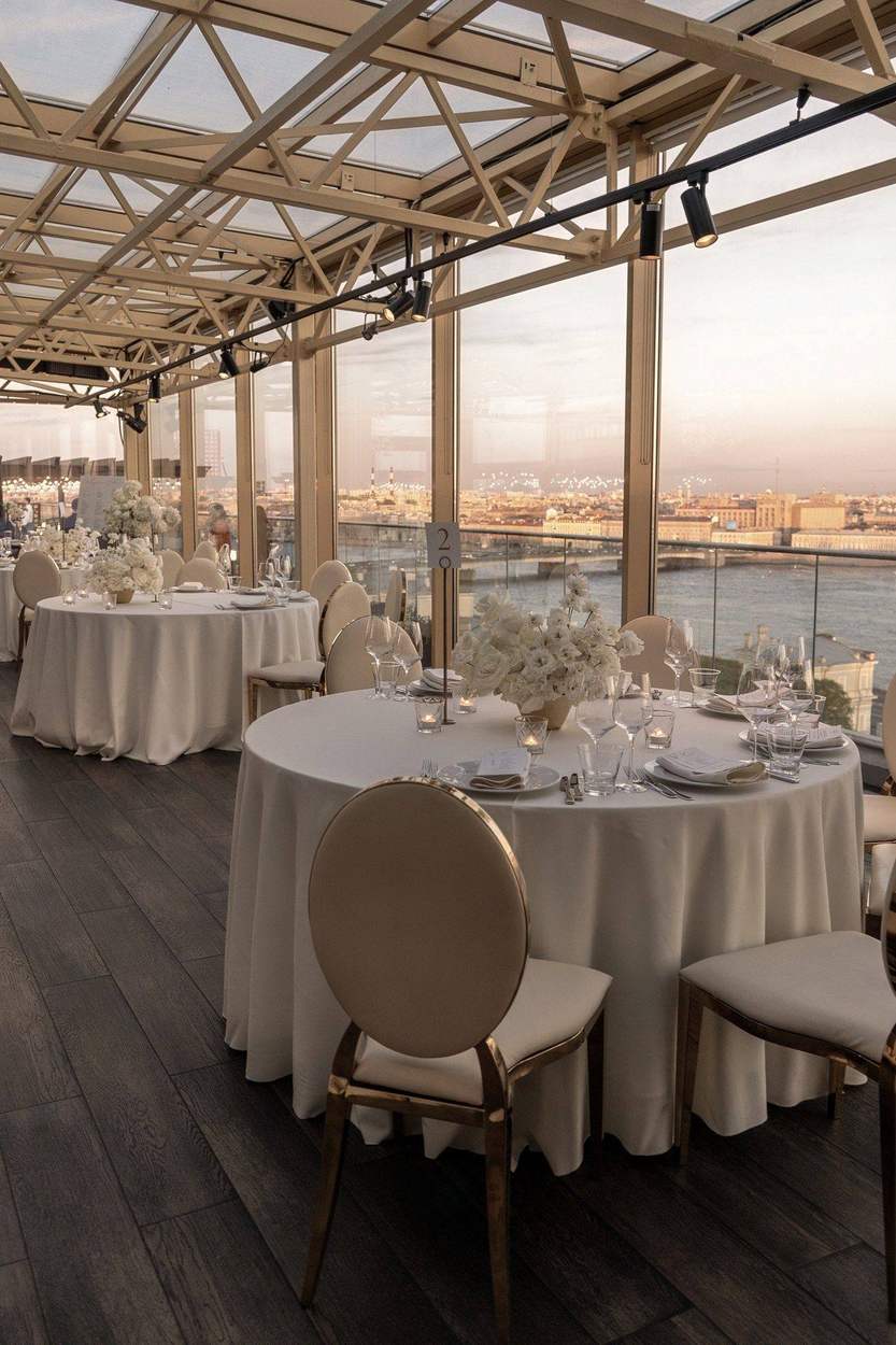 Washington DC photographer wedding venue details: round reception tables with white floral centerpieces in a glass-roofed room overlooking the river at sunset.