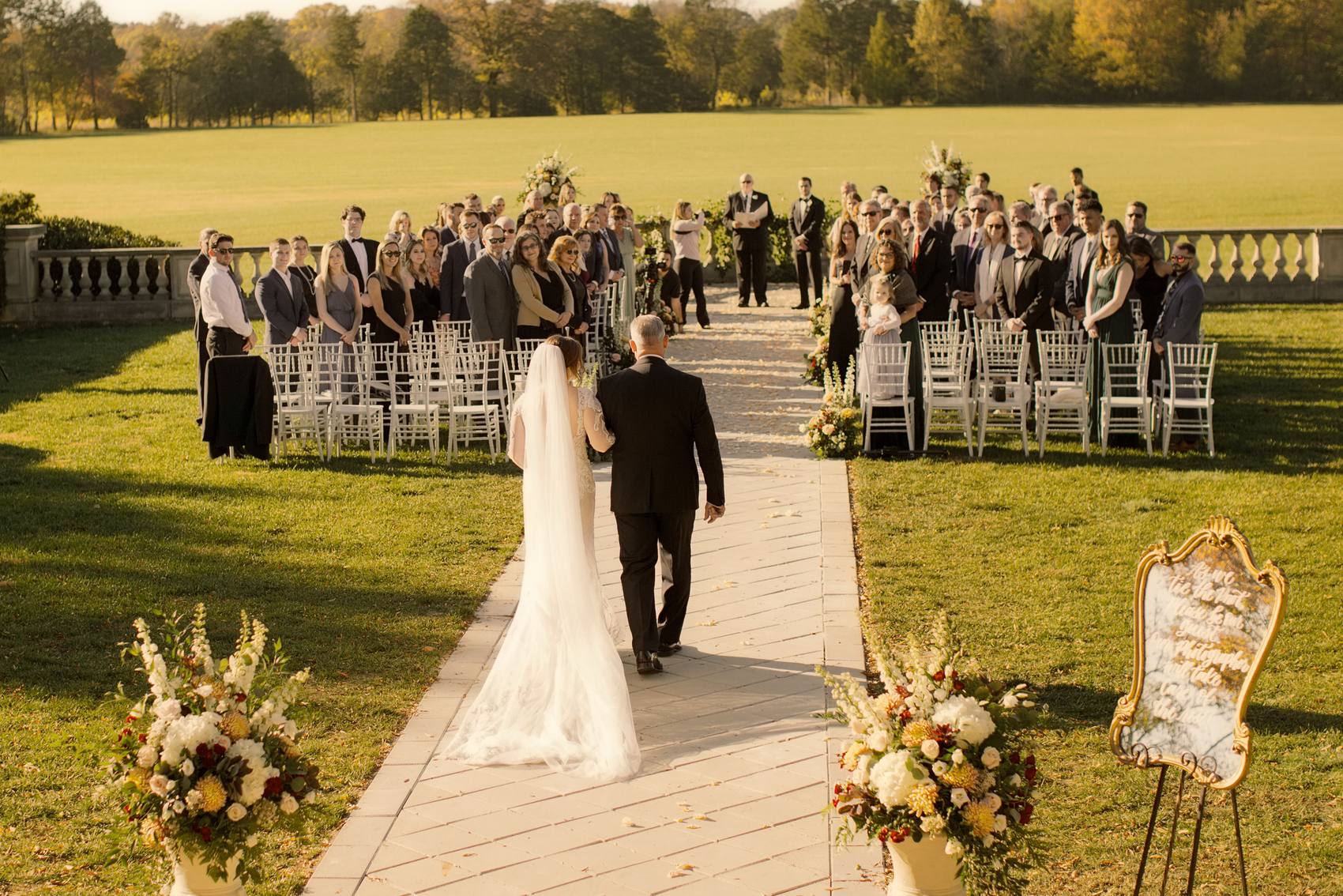 View from behind of the bride in a long veil walking up a stone aisle on the arm of a man in a black suit toward standing guests and an officiant, flower urns beside the path and a wide lawn beyond