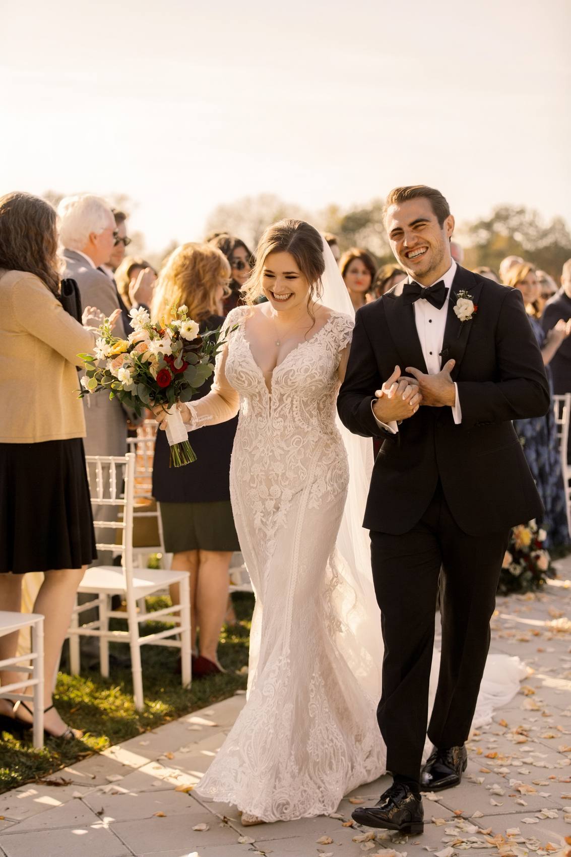 Bride holding her bouquet and groom arm in arm, both laughing as they walk up the petal-strewn aisle past clapping guests