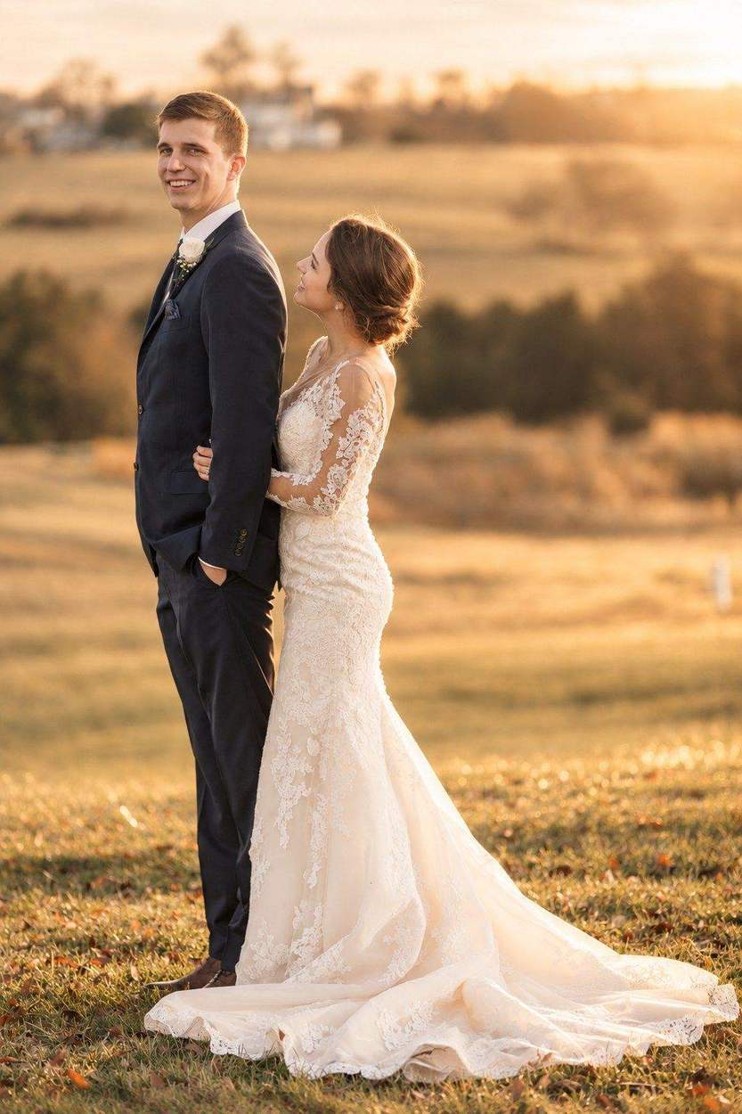 Washington DC photographer autumn wedding portrait: a groom smiling over his shoulder as his bride in a lace gown embraces him from behind, in golden sunset light over rolling fields.